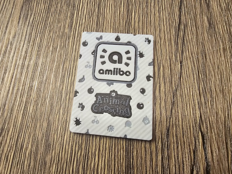 May include: Amiibo card featuring the Animal Crossing logo. The card has a white background with a pattern of small black icons. The Amiibo logo is in a white square with a black border. The Animal Crossing logo is in a black banner.