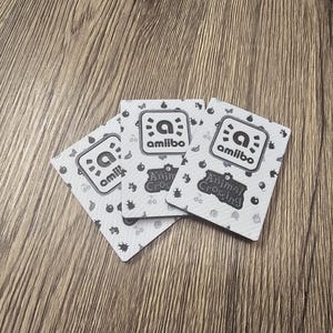 May include: Three Animal Crossing amiibo cards are shown on a wooden surface. Each card displays the amiibo logo and the words "Animal Crossing". The cards have a white background with a pattern of small icons.