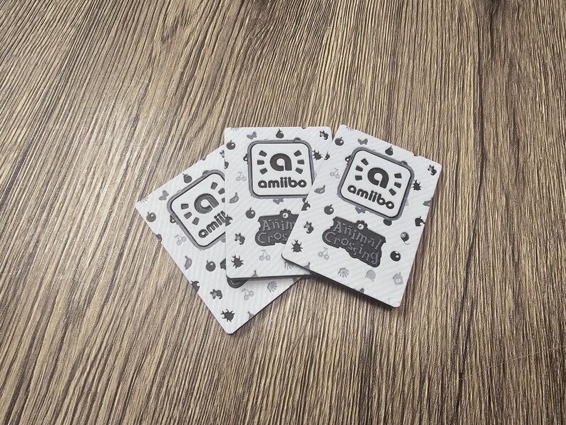 May include: Three Animal Crossing amiibo cards are shown on a wooden surface. Each card displays the amiibo logo and the words "Animal Crossing". The cards have a white background with a pattern of small icons.