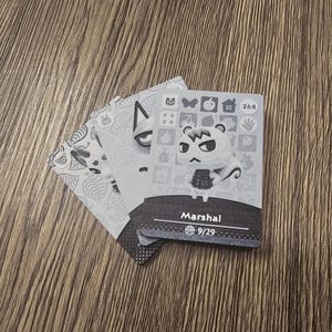 May include: Three Animal Crossing character cards are displayed on a wooden surface. The top card features a character named Marshal, with a white face and black eyes. The cards are grayscale with decorative patterns.