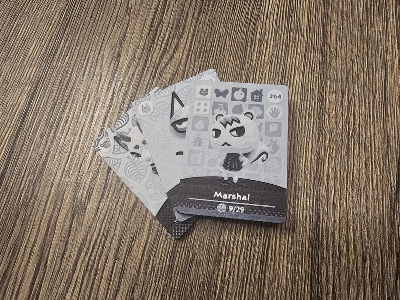 May include: Three Animal Crossing character cards are displayed on a wooden surface. The top card features a character named Marshal, with a white face and black eyes. The cards are grayscale with decorative patterns.