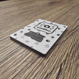 May include: A white Animal Crossing Amiibo card with a black border. The card features the Amiibo logo and the Animal Crossing logo. The card is decorated with small, grey icons. The card is resting on a wooden surface.