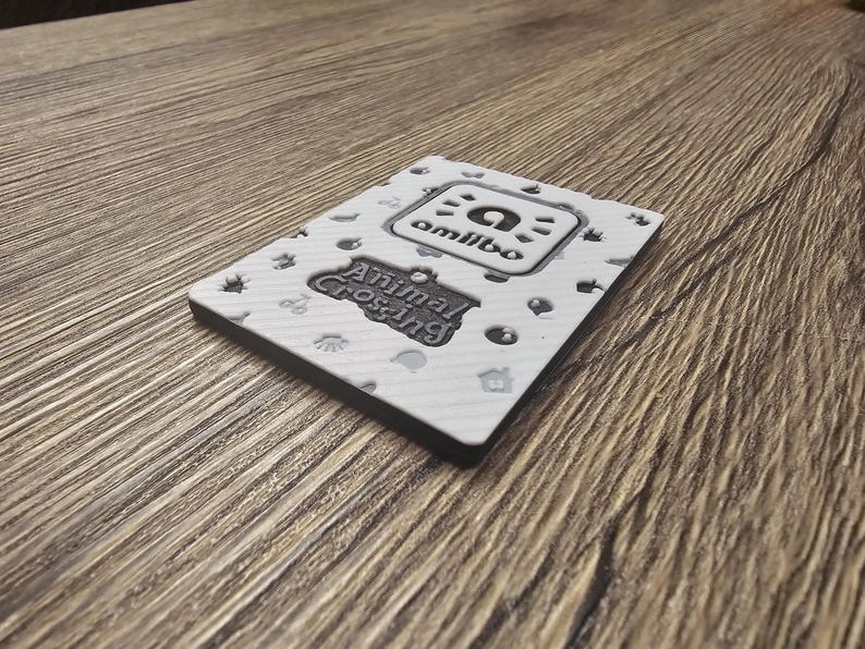 May include: A white Animal Crossing Amiibo card with a black border. The card features the Amiibo logo and the Animal Crossing logo. The card is decorated with small, grey icons. The card is resting on a wooden surface.