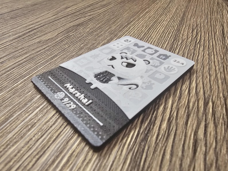 May include: A grey and white card with the name "Marshal" and the number "9/29". The card features a cartoon character and various icons. The surface has a textured finish.