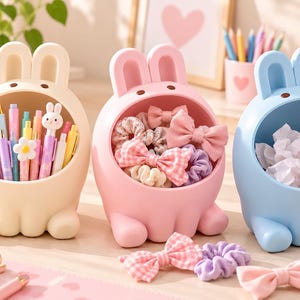May include: Three rabbit-shaped desk organizers in cream, pink, and blue. The cream organizer holds various pens and pencils. The pink organizer contains fabric hair bows and scrunchies. The blue organizer holds crumpled paper.
