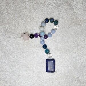 May include: A handmade beaded necklace featuring a variety of colorful gemstones. The necklace includes a rose quartz bead, amethyst beads, and a dark blue rectangular pendant. The beads are strung on a white cord.