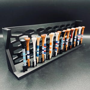 May include: A black golf tee holder with a honeycomb design, holding a row of golf tees. The tees are wooden with various painted designs, including stripes and solid colors. Some tees have the text "Jeremy Head".