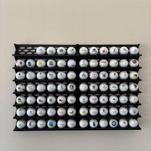 May include: A black wall-mounted display rack with a honeycomb-patterned back panel, featuring eight horizontal shelves. The rack holds a collection of white golf balls, each printed with various colorful logos, emblems, and text designs.