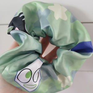 Puede incluir: Una scrunchie de satén verde con una cara de dibujos animados y un estampado floral. La scrunchie presenta una cara blanca con un ojo verde, cabello negro y un contorno azul. El fondo tiene un diseño floral verde claro, azul y blanco.