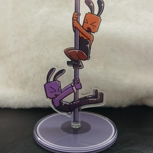 May include: An acrylic standee featuring two cartoon rabbit-like characters with block-shaped heads. One orange character climbs a vertical pole, while a purple character hangs from the same pole. Both figures wear dark clothing and bunny ears.