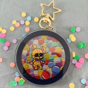 May include: A clear, round keychain with a gold star-shaped clasp. Inside, a cartoon character is surrounded by colorful confetti. The keychain has a dark blue border and is decorated with a gold-colored star-shaped clasp.