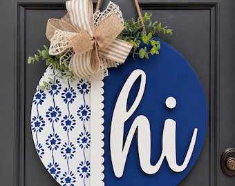 Könnte beinhalten: Runder blau-weißer Holztürschild mit dem Wort "hi" in weißer Schrift. Das Schild ist mit einem Muster, einer dekorativen Schleife und Grün geschmückt. Es hängt an einem Juteseil.