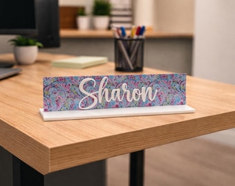 Könnte beinhalten: Ein Schreibtisch-Namensschild mit dem Namen "Sharon" in weißer Schrift. Das Namensschild hat ein farbenfrohes Blumenmuster in Blau-, Rosa- und Lilatönen. Es steht auf einem weißen Sockel, der auf einem Holzschreibtisch ausgestellt ist.
