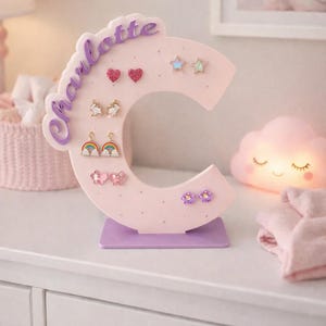 May include: A pink letter "C" shaped earring holder with the name "Charlotte" in cursive. The holder displays various earrings, including hearts, stars, unicorns, rainbows, butterflies, and flowers. The base is purple.