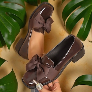 May include: Brown patent leather loafers with a square toe and a large bow detail. The shoes have a low heel and a glossy finish. The image is set against a backdrop of green leaves and a light brown surface.