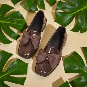 May include: A pair of brown patent leather loafers with large bow accents. The shoes have a square toe and a low heel. The background features green tropical leaves on a cream-colored surface.