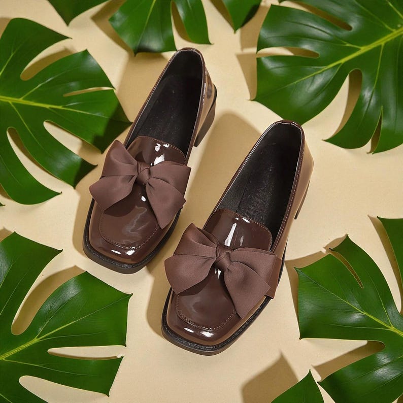 May include: A pair of brown patent leather loafers with large bow accents. The shoes have a square toe and a low heel. The background features green tropical leaves on a cream-colored surface.