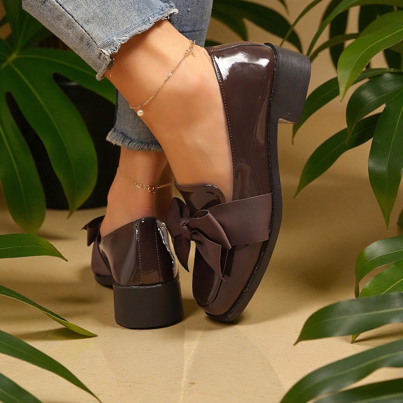 May include: Brown patent leather loafers with a bow detail. The shoes have a low block heel and a glossy finish. The bow is made of a matching brown fabric. The shoes are worn with denim jeans.