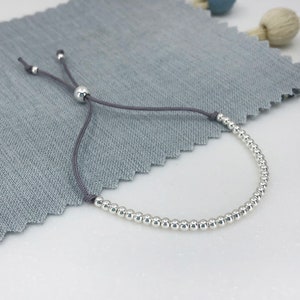 May include: A silver bead bracelet with a grey adjustable cord. The bracelet is made of small, round beads and has a simple, minimalist design.
