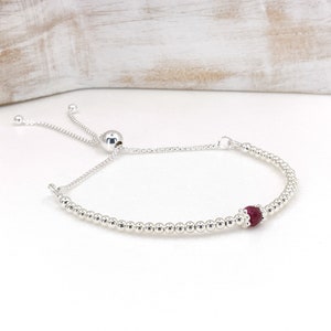 May include: A silver beaded bracelet with a red gemstone accent. The bracelet has a sliding clasp for adjustable sizing.