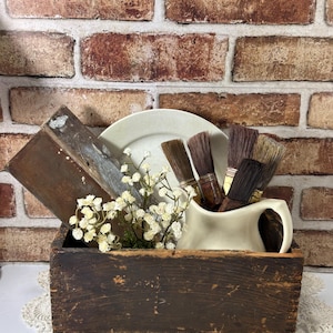 May include: A rustic wooden box filled with vintage paint brushes, a white ceramic pitcher, a white plate, and small white flowers. The box sits on a white lace doily against a brick wall background.