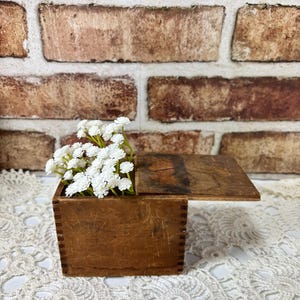 May include: A small, square, brown wooden box with visible finger joints at the corners and a sliding lid. The box contains a cluster of small, white artificial flowers with delicate petals, creating a contrast against the wood grain.