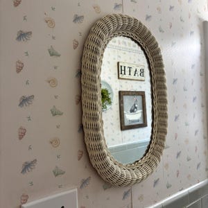 May include: An oval wall mirror featuring a light beige woven wicker frame. The mirror surface reflects a room interior with a sign that reads BATH and a framed picture of a creature in a bathtub, set against wallpaper with a seashell pattern.