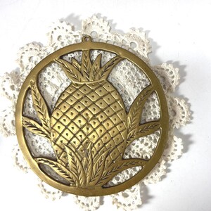 May include: A gold-colored metal pineapple trivet with a circular frame. The pineapple design is detailed with textured diamond shapes and leafy accents. The trivet is placed on a white, lacy doily.