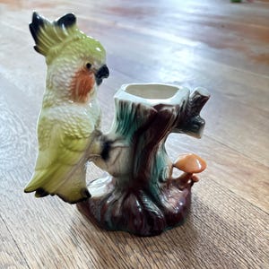 May include: A ceramic figurine featuring a yellow and white cockatoo perched on a brown tree stump. The stump has a hollow top and a small orange mushroom at its base. The bird has a crest, a black beak, and orange cheek patches on its face.