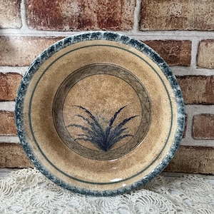 May include: A round ceramic bowl featuring a tan base with a fine crackle glaze finish. The bowl has a dark blue, mottled rim and a central circular band enclosing a dark blue, stylized botanical illustration of a grass-like plant design.