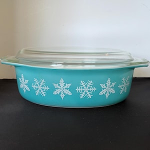 May include: A turquoise oval casserole dish with a clear glass lid. The exterior of the dish features a repeating pattern of white snowflakes encircling the sides. The dish has integrated handles on both ends and a smooth, glossy finish.