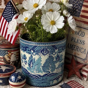 May include: A cylindrical metal container featuring a blue and white willow pattern, filled with white cosmos flowers. The scene includes a small American flag, a round patriotic ball, a metal tin with an eagle, and a sign reading GOD BLESS AMERICA.