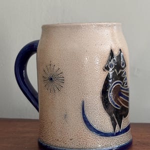 May include: A beige ceramic mug with a textured surface, featuring a dark blue handle and base. The mug is decorated with a stylized black and blue owl graphic on the side, accompanied by a small, etched starburst pattern to the left of the owl.