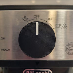May include: Close-up of a stainless steel espresso machine control panel. A black dial with a white indicator points to the "OFF" position. The panel has icons for steam and coffee, and the brand name "DeLonghi" is visible.
