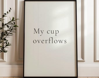 May include: A framed print with the text "My cup overflows" in a minimalist style. The black framed art piece is displayed against a white wall and a wooden floor, creating a clean and modern aesthetic. The artwork is suitable for home decor.