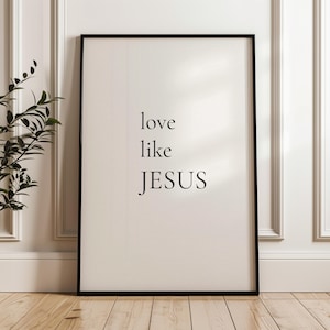 Puede incluir: Una impresión enmarcada con las palabras "love like JESUS" en una fuente negra minimalista. La impresión está sobre un fondo crema y enmarcada en un borde negro. La imagen está apoyada contra una pared blanca.