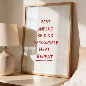May include: A framed print with the words "REST, UNPLUG, BE KIND, TO YOURSELF, HEAL, REPEAT" in red lettering. The print is in a light wood frame and is displayed on a nightstand next to a lamp.