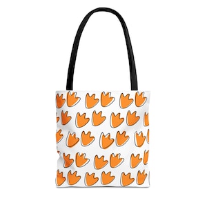May include: A white tote bag featuring a repeating pattern of orange duck footprints with black outlines. The bag has two black fabric handles attached to the top, providing a sturdy grip for carrying items in this functional accessory.