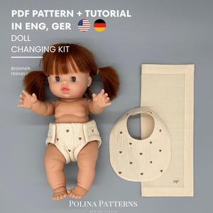 Könnte beinhalten: Ein Puppen-Wickelset PDF-Schnittmuster und Anleitung mit einer Puppe mit rotbraunen Zöpfen, die eine cremefarbene Windel mit braunen Herzen trägt, dazu ein passendes cremefarbenes Lätzchen mit braunen Herzen und ein rechteckiges Tuch mit MP.