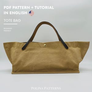 Könnte beinhalten: Eine beigefarbene Tragetasche mit schwarzen Griffen und einem goldenen Knopfverschluss. Die Tasche besteht aus einem strukturierten Material und trägt den weißen Text "PDF PATTERN + TUTORIAL IN ENGLISH" und "TOTE BAG". Die Worte "BEGINNER-FRIENDLY" sind ebenfalls sichtbar.