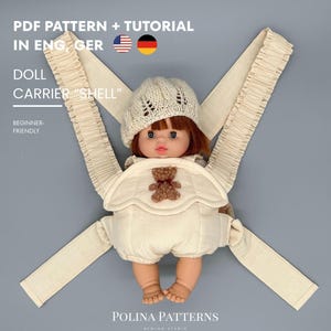 Op de afbeelding: Een pop in een crèmekleurige outfit en gebreide hoed zit in een crèmekleurige poppendraagzak met gerimpelde bandjes. De afbeelding bevat tekst: "PDF PATTERN + TUTORIAL" en "DOLL CARRIER 'SHELL'". De draagzak is geschikt voor beginners.
