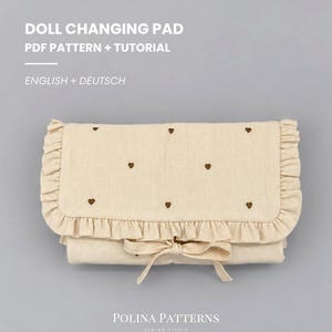 Könnte beinhalten: Ein PDF-Schnittmuster und eine Anleitung für eine Puppenwickelunterlage von Polina Patterns. Der beigefarbene Stoff hat kleine braune Herzstickereien, einen Rüschenrand und eine Stoffschleife. Der Text lautet DOLL CHANGING PAD PDF PATTERN + TUTORIAL.