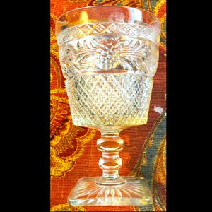May include: A clear, ornate glass goblet with a square base and a detailed stem. The bowl of the glass features diamond and floral patterns. The rim of the glass has a decorative border. The goblet is likely vintage or antique.