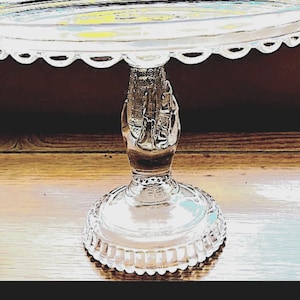 May include: A clear glass cake stand with a scalloped edge. The stand has a detailed base and a central support featuring a sculpted design. The stand is on a wooden surface.