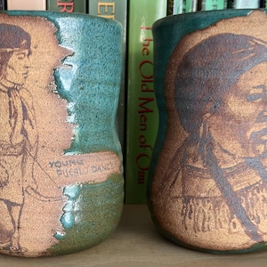 May include: Two ceramic mugs with a teal glaze and textured brown panels. The left mug features a figure in profile with the text "YOUNG PUEBLO DANCER". The right mug displays a portrait of a person with braided hair. Both mugs have a cylindrical shape.