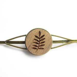 Può includere: Un fermaglio per capelli con un elemento centrale rotondo in legno marrone chiaro con un disegno a foglia intagliata. Il fermaglio ha una montatura in metallo color bronzo. Il design è semplice ed elegante, adatto a varie acconciature.