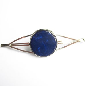 Può includere: Un fermaglio per capelli in argento con un cabochon rotondo in resina blu. Il cabochon ha un effetto marmoreo.