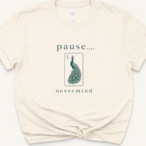 May include: Cream-colored t-shirt with the words "pause...." and "nevermind" in dark green. A peacock illustration is centered between the words. The peacock is in a rectangular frame with the number "1" at the top.