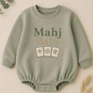 May include: Sage green baby onesie with long sleeves and a round neckline. The front features the text "Mahj Baby" in shades of green and tan, along with three Mahjong tile graphics. The onesie has snap closures at the bottom.