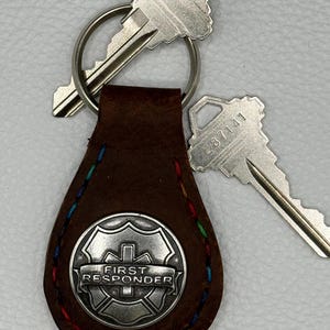 May include: A brown leather keychain with a silver emblem that reads "FIRST RESPONDER." The keychain has a silver key ring with two silver keys attached. The edges of the leather are stitched with colorful thread.